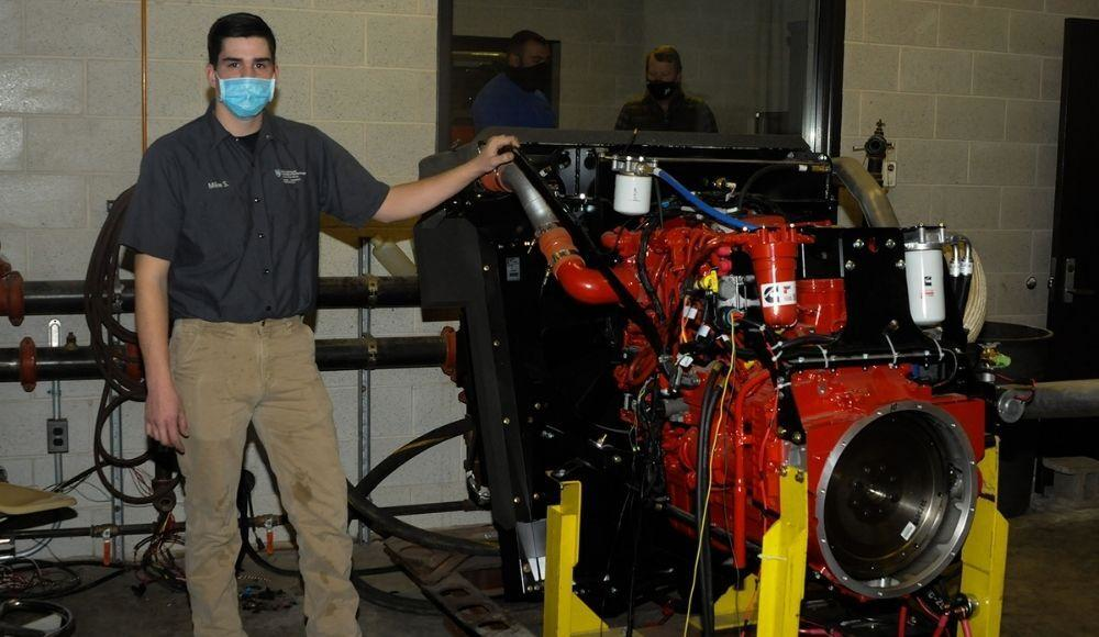 Michael J. Sormilic, an online power generation student from Southbury, Conn., who has spent much of the Fall 2020 semester learning the ins and outs of the equipment, stands alongside a Cummins crane engine donated to Pennsylvania College of Technology by Terex Corp. Behind him, in the control room of the dyno lab, are John D. Motto (left), diesel equipment technology instructor, and Chris Macdonald, assistant director of corporate relations.