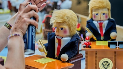 A woman uses hairspray to fixe the hair on a nutcracker titled 'Resolute Desk of the President,' featuring a seated figure signing a 'presidential proclamation,' at the Steinbach-Volkskunst nutcracker factory in Marienberg, Germany, Tuesday, April 15, 2025.