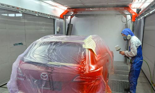 A painter works in a Symach equipped booth. A RoboDry Plus, one of the company's drying robots, is visible in the background.
