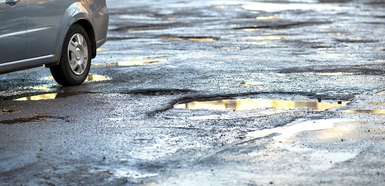 Close up of car wheel on a road in very bad condition with big potholes full of dirty rain water pools.