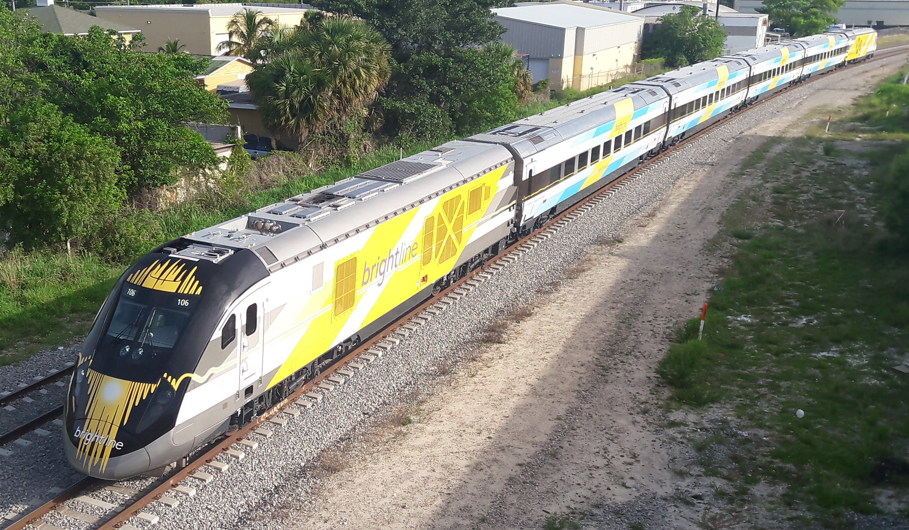 Brightline passenger train traveling on tracks through residential area with trees and buildings