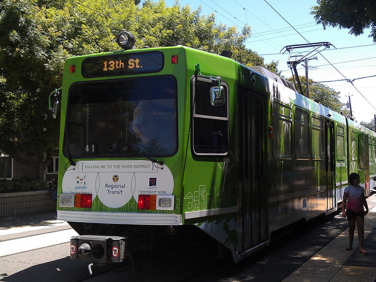 Bright green and black light rail tram marked '13th St' traveling on tree-lined street with overhead power lines