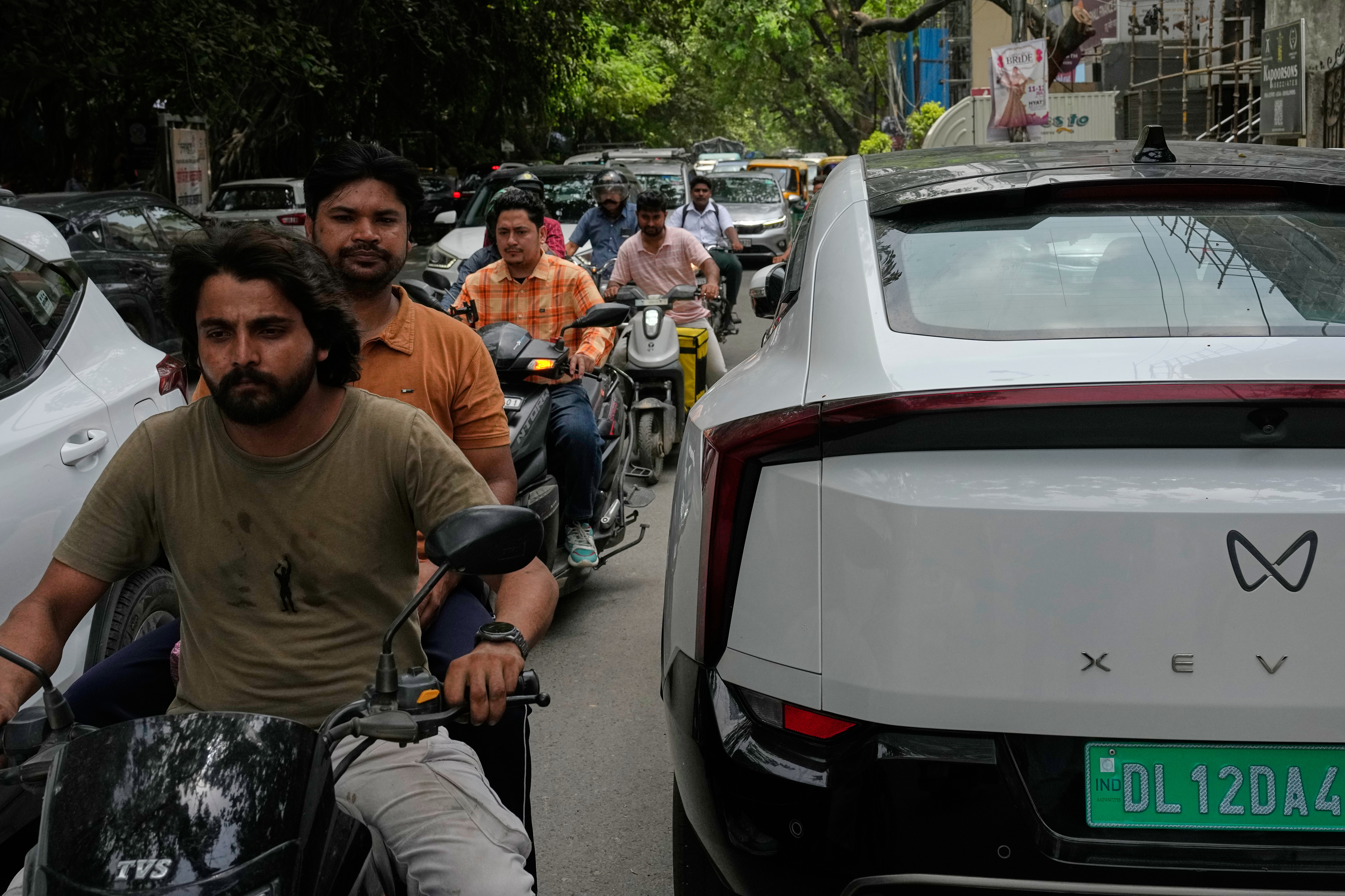 Two wheelers drive past an electric vehicle parked along a street in New Delhi, India, Monday, July 13, 2026.